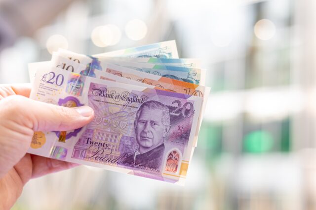 Close-up of a hand holding British pound banknotes (£5, £10, £20), featuring King Charles III, representing calculating investment and providing bad financial advice.