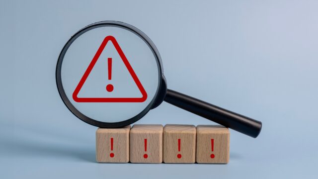 A magnifying glass is placed on top of a stack of wooden blocks with red exclamation points on them. Concept of urgency and danger, as the red exclamation points