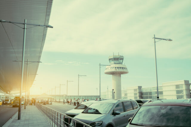 Parking lot and road near modern contemporary airport terminal in front of air traffic control tower, with many passengers and staff passing in distance.
