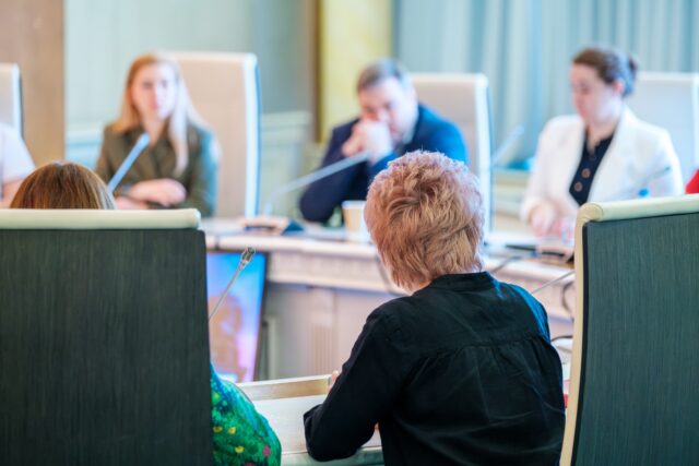 Business people sitting at round table with colleagues using microphones during conference in spacious hall.