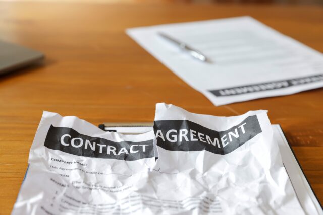 A crumpled contract agreement on a wooden desk, symbolizing a failed deal or negotiation. Close-up of a damaged contract on a work desk, representing a dispute or cancellation.